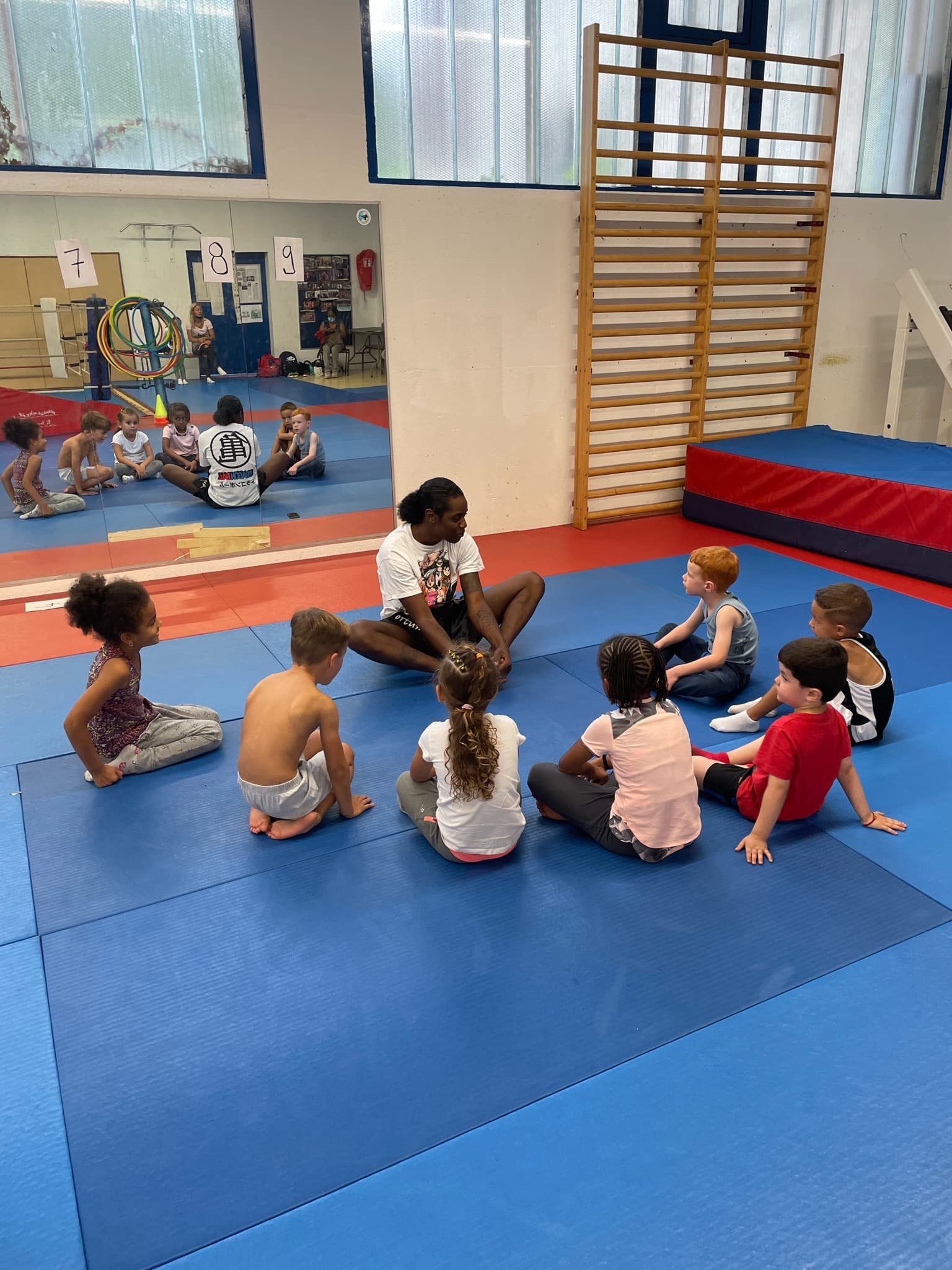 Un coach assis en tailleur explique un exercice à un groupe de jeunes enfants sur les tatamis.